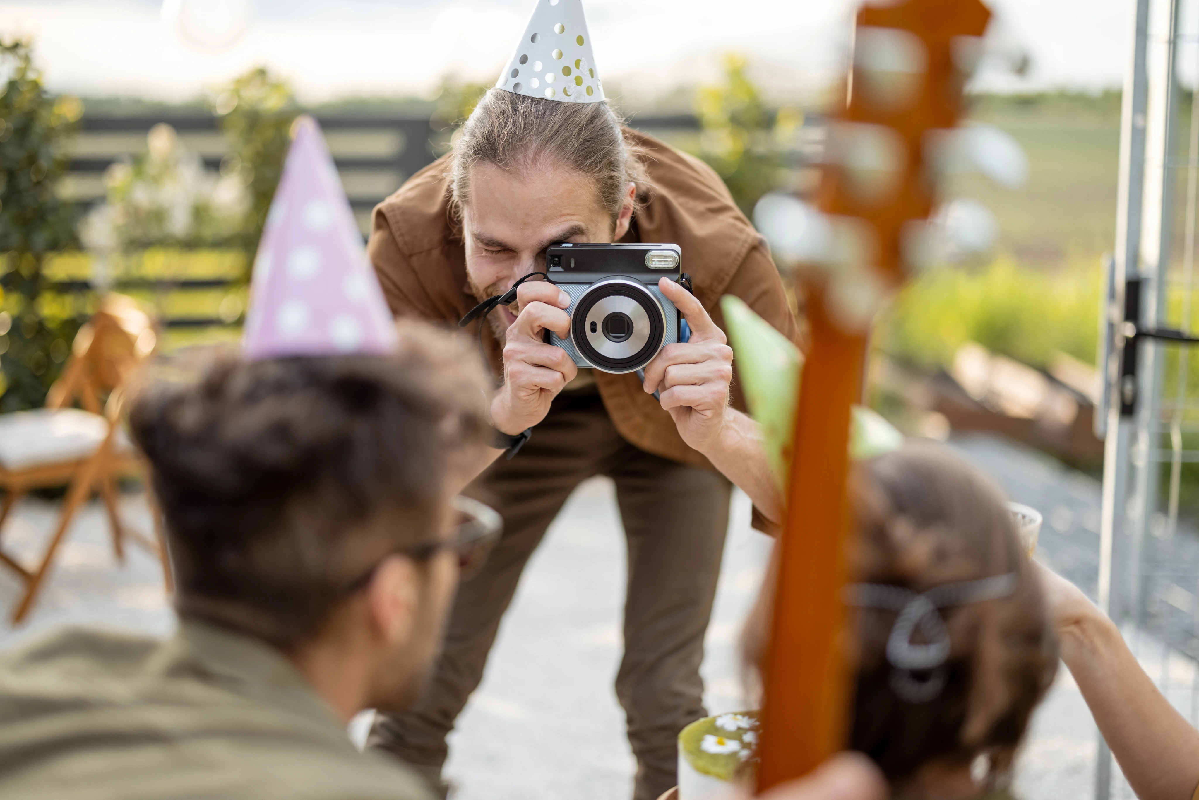 A photographer wearing a party hat and brown jacket leans over to take a photo of guests at an outdoor celebration with party decorations visible