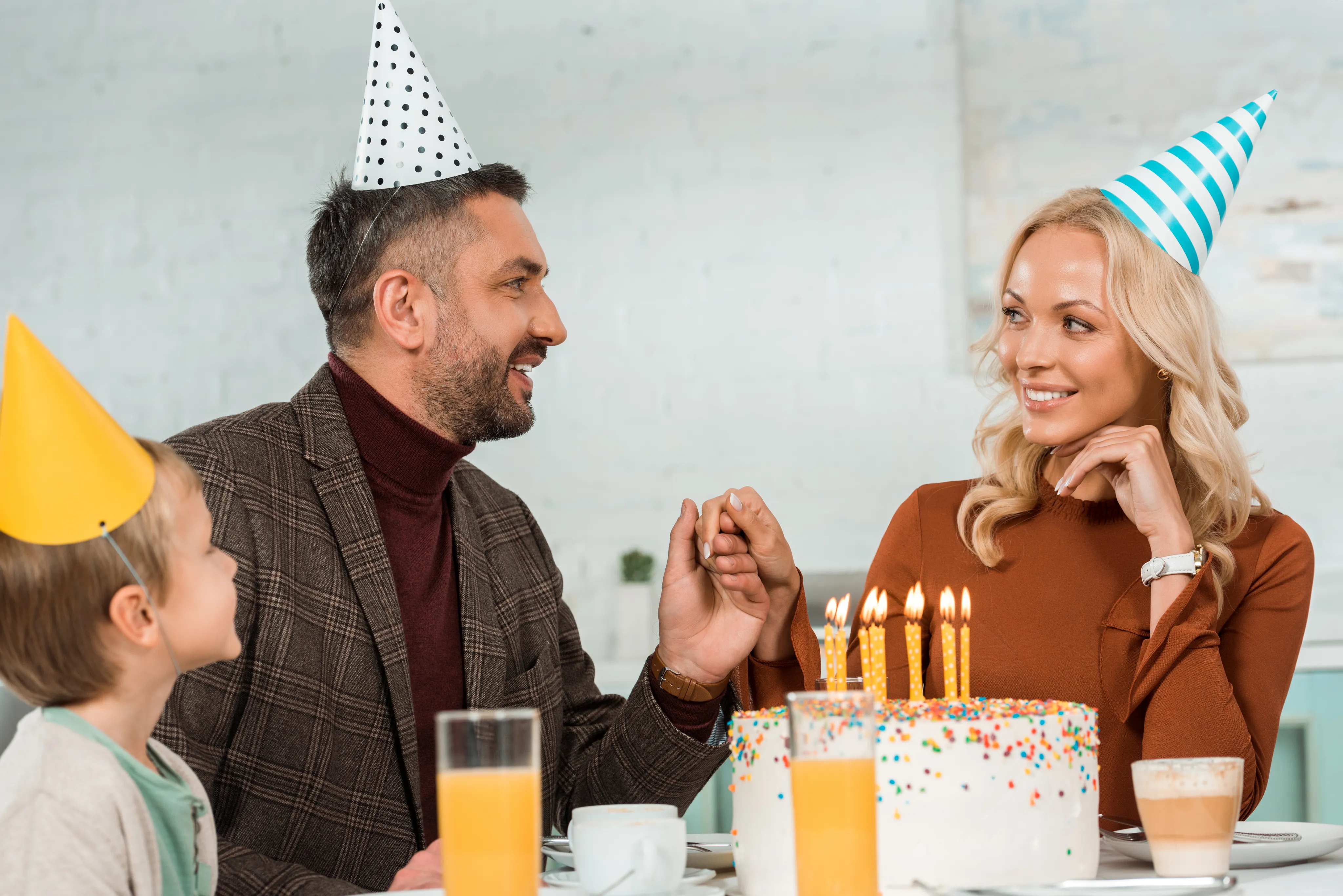 A family birthday celebration with three people wearing party hats - a child, a man in a brown turtleneck and plaid jacket, and a woman in a brown sweater - gathered around a white birthday cake with lit candles. Orange juice and coffee are on the table.