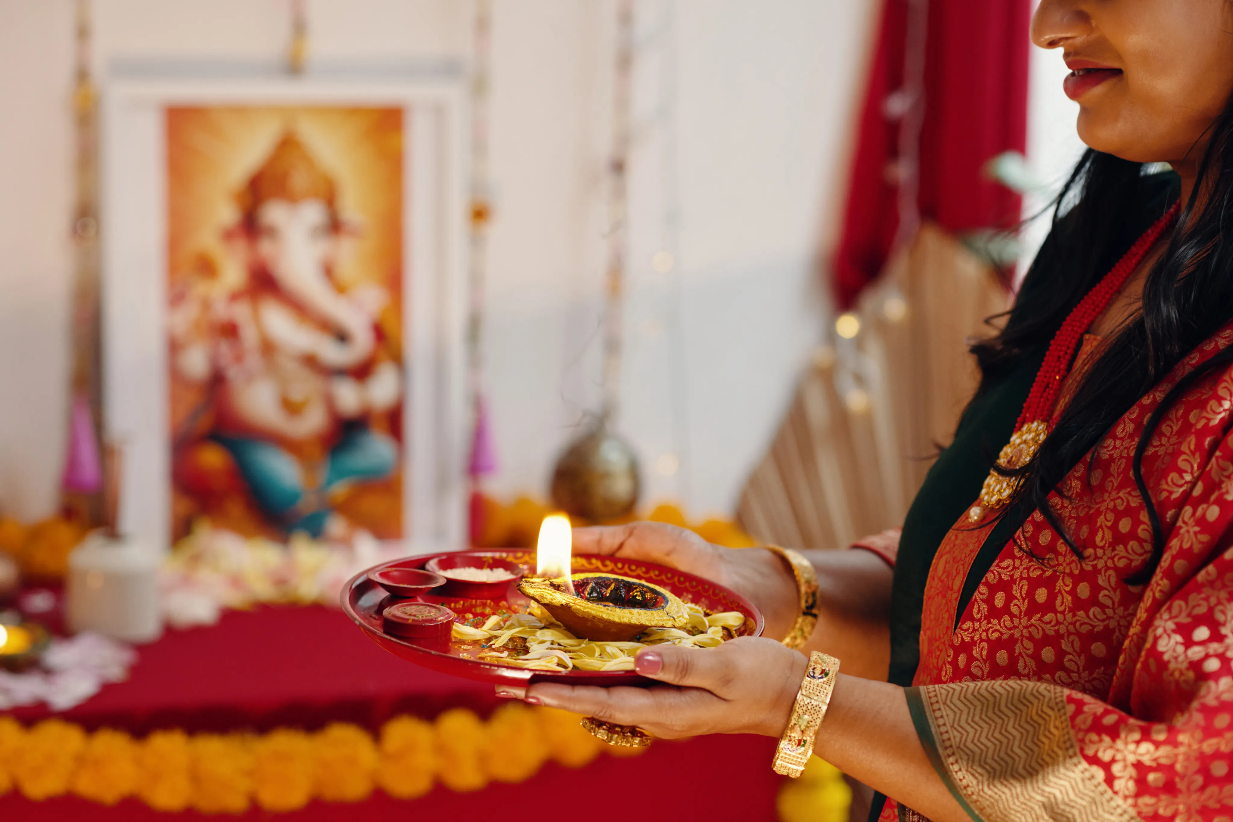 A religious ceremony setting with a Ganesha portrait in the background. Someone wearing traditional Indian attire with gold jewelry holds a decorated thali (plate) with a lit diya (oil lamp) and offerings against a red and gold backdrop.