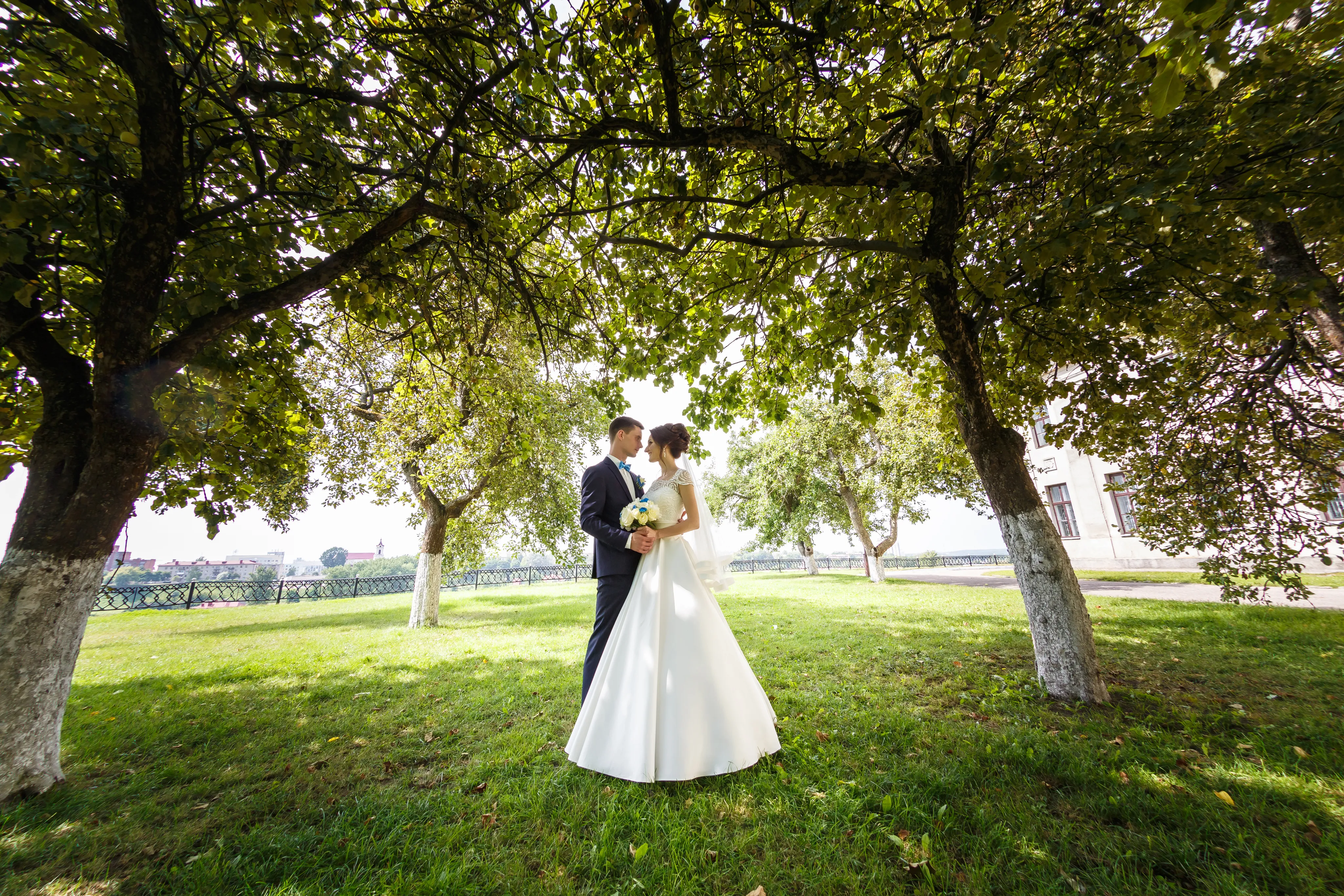 A wedding photograph taken outdoors showing a bride in a white ball gown and groom in a black tuxedo standing close together under a natural arch created by large trees with green leaves. A manicured lawn and ornate fence are visible in the background.