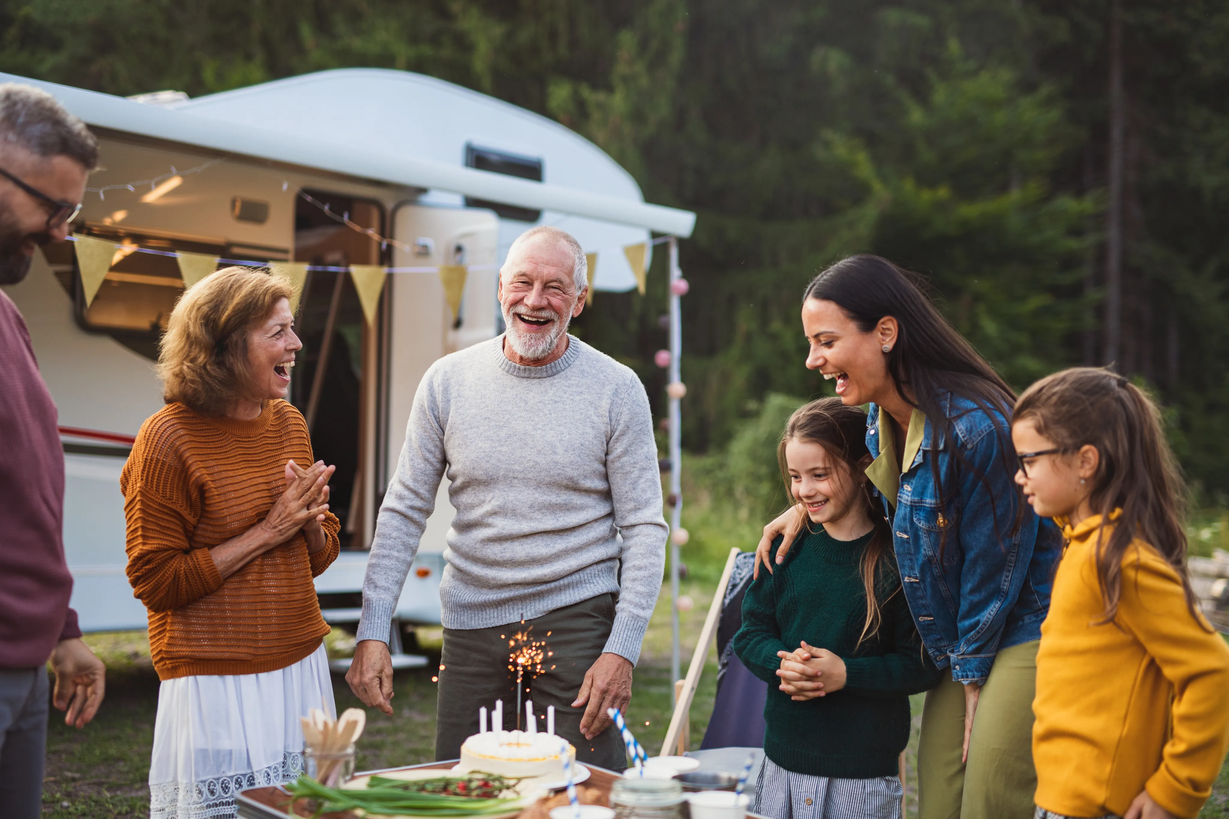 A multi-generational family gathering outdoors by an RV. People of various ages including children and seniors are laughing together around a birthday cake with sparklers. They are wearing casual sweaters and the setting appears to be a campground or RV park.