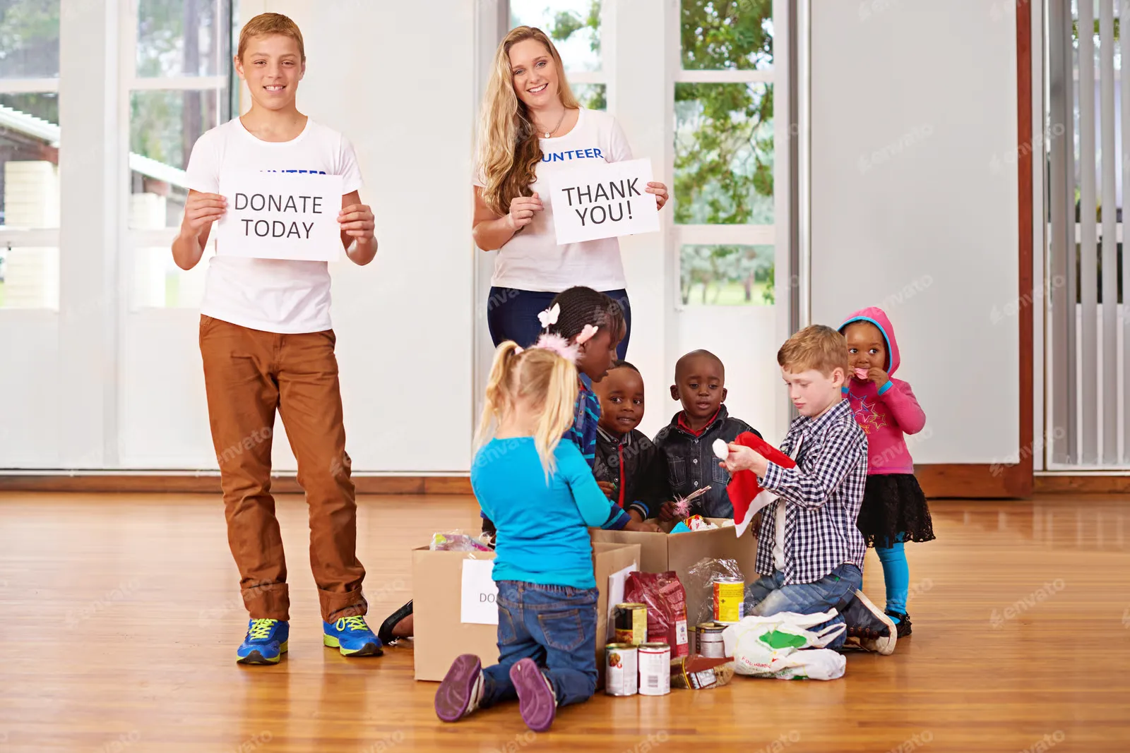 A charity donation event with young volunteers in white VOLUNTEER t-shirts holding signs saying DONATE TODAY and THANK YOU! while children sort through donation boxes containing food items and supplies on a hardwood floor.