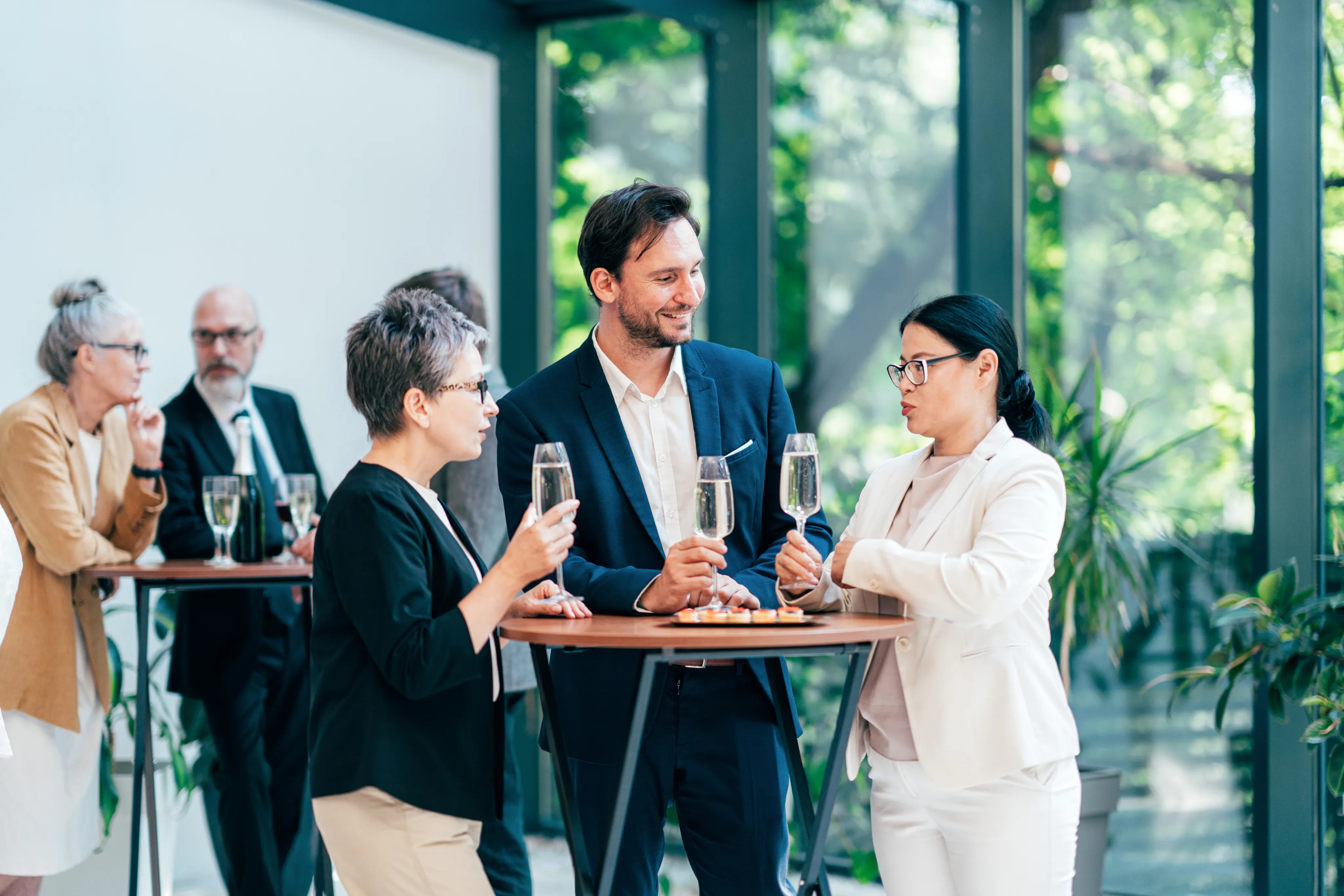 A professional networking event in a modern space with floor-to-ceiling windows and plants. Three people in business attire are having a conversation while holding champagne glasses around a high table, with two more people visible in the background.